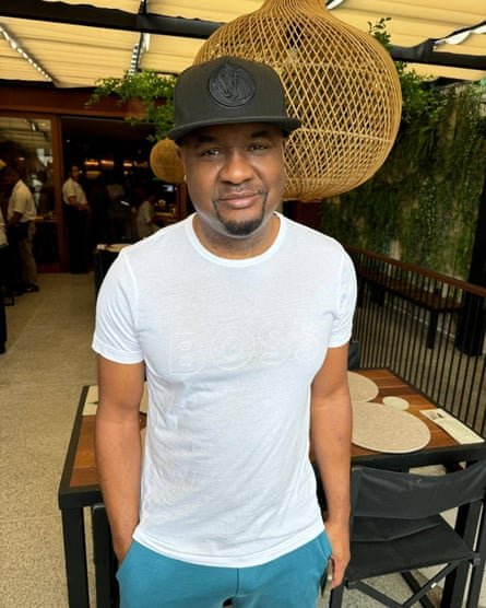 A man in a cap and white T-shirt smiles with his right hand in his pocket